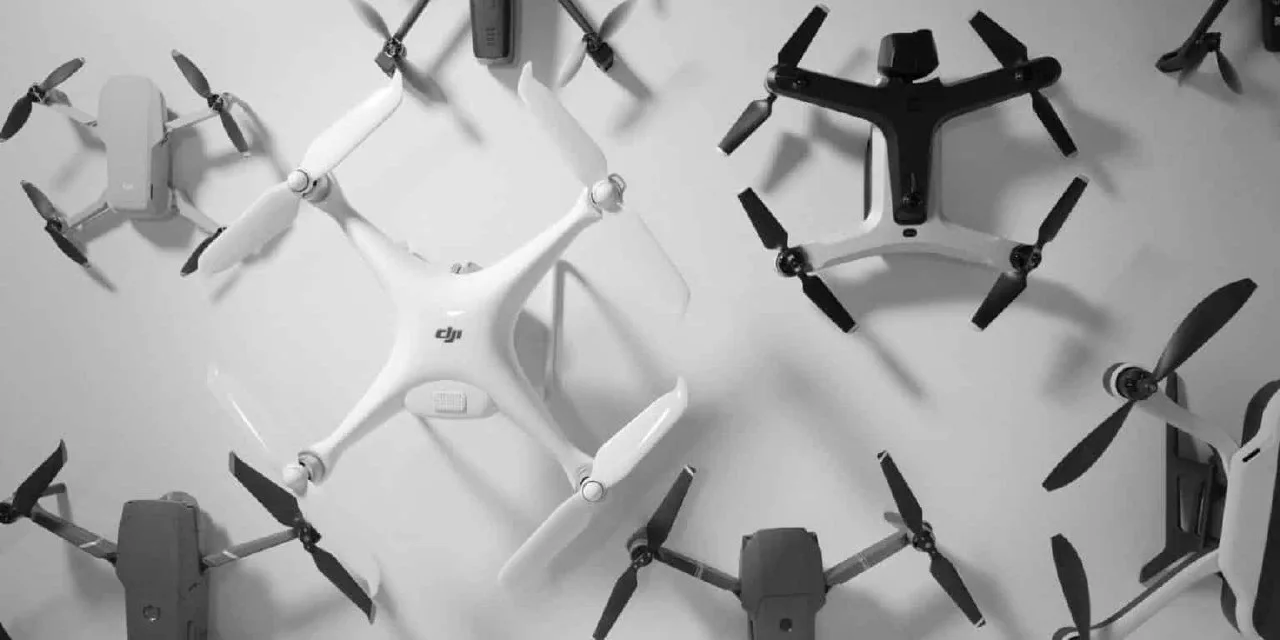 Overhead black-and-white view of a DJI Phantom quadcopter surrounded by folding DJI Mavic drones and other multirotors arranged on a white surface