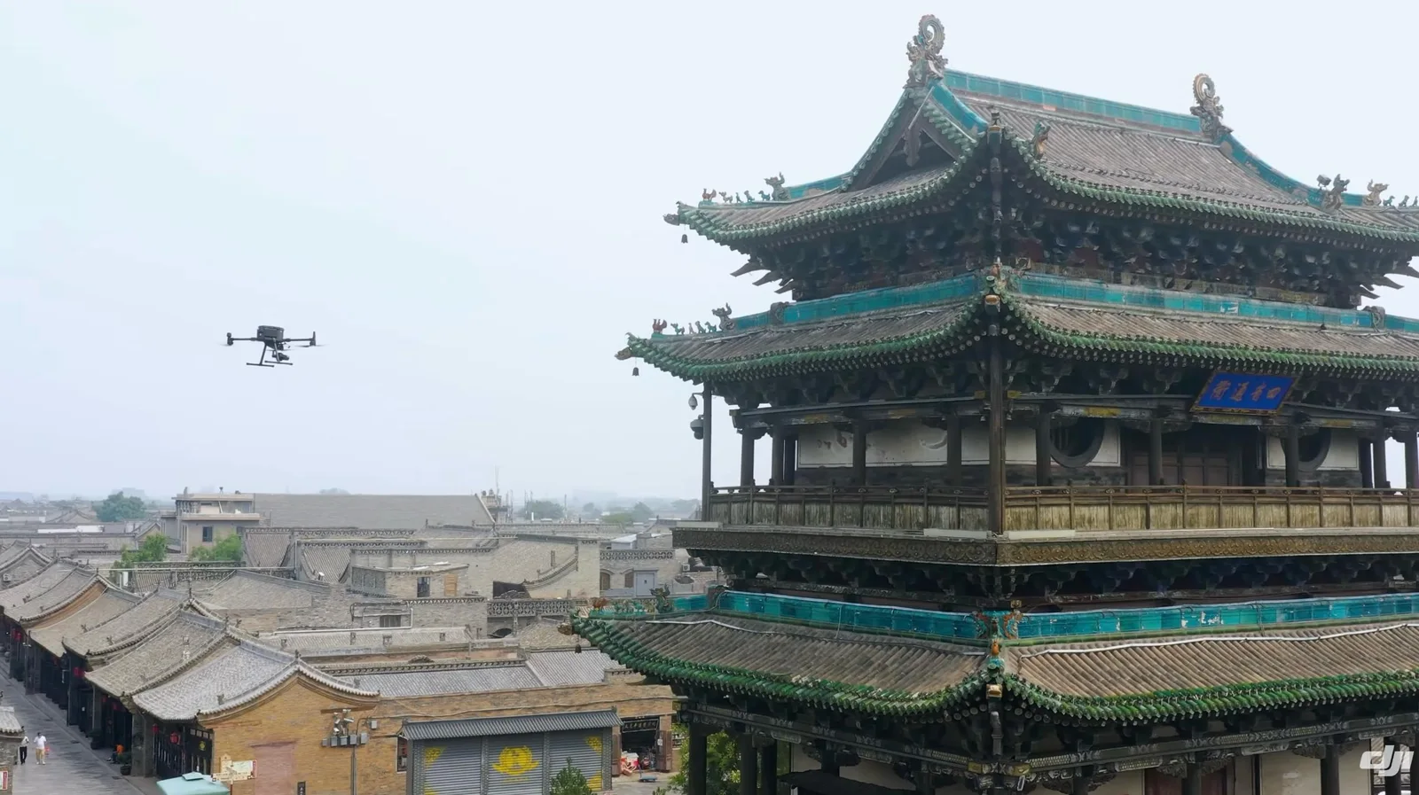 A DJI enterprise drone hovers beside the ornate green-tiled eaves of a multi-story wooden Chinese heritage tower, with the gray-roofed old town spread out below.