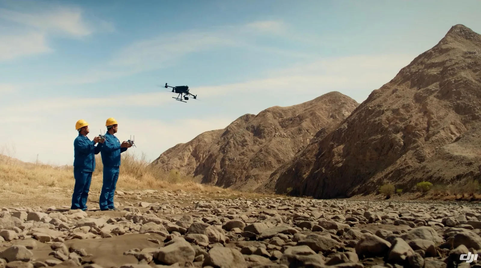 Two surveyors in blue uniforms and yellow hard hats pilot a DJI Matrice drone over a dry rocky riverbed beside steep arid mountains