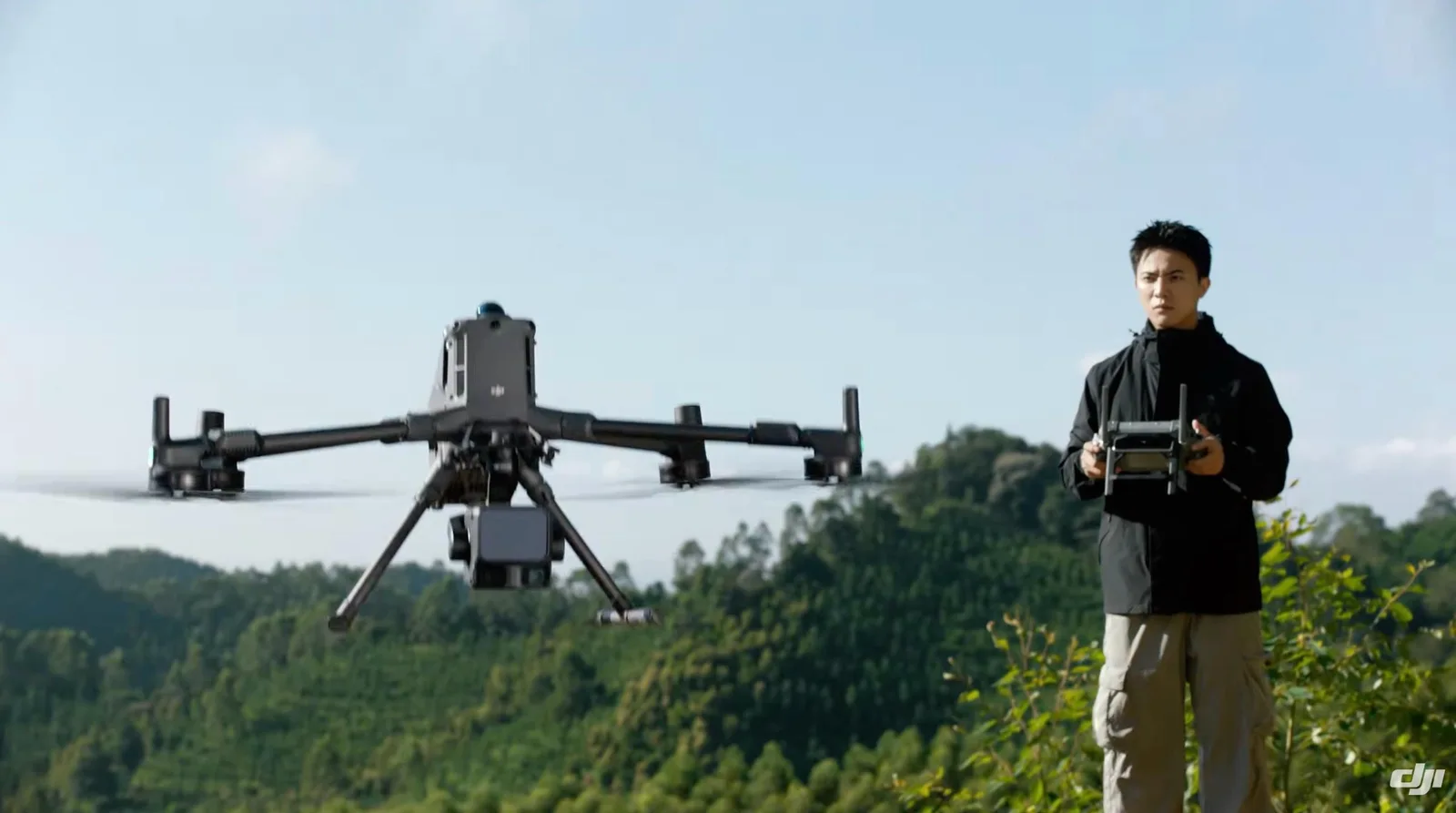 A pilot in a black jacket flies a DJI Matrice enterprise drone with a gimbal payload over forested hills
