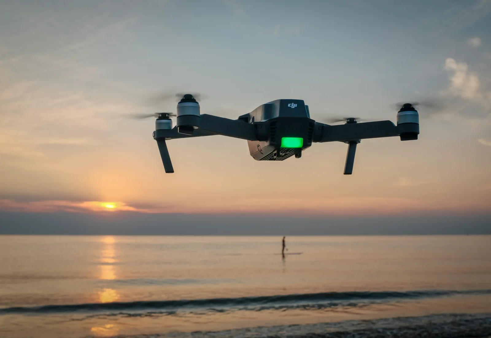 DJI Mavic Pro drone hovering above a calm beach at sunset with a paddleboarder on the water in the background