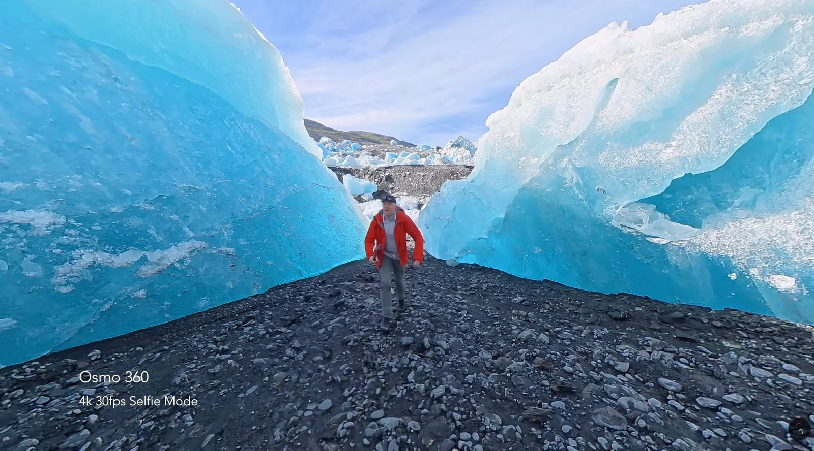 DJI Osmo 360 selfie-mode frame showing a hiker in an orange jacket walking between towering blue glacial ice walls