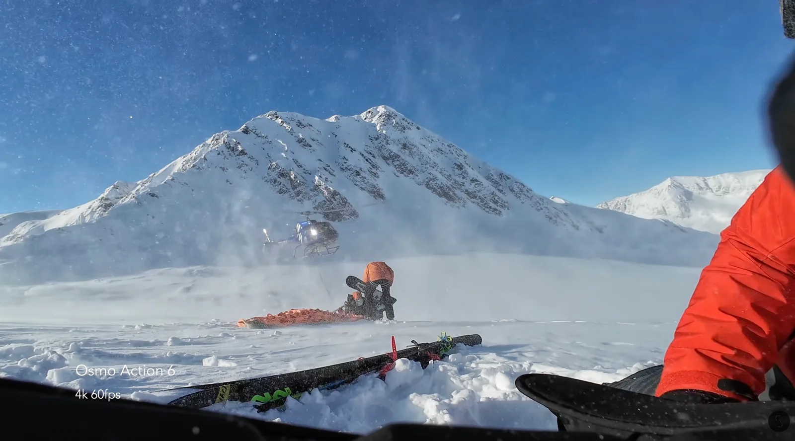 DJI Osmo Action 6 4K 60fps frame showing a helicopter hovering over a snow-covered mountain bowl with skis and a rider in an orange jacket in the foreground
