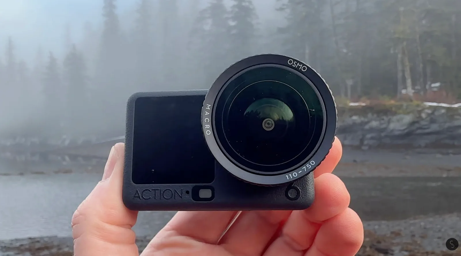 Hand holding a DJI Osmo Action camera fitted with a screw-on macro lens, with a foggy pine forest and rocky shoreline behind it