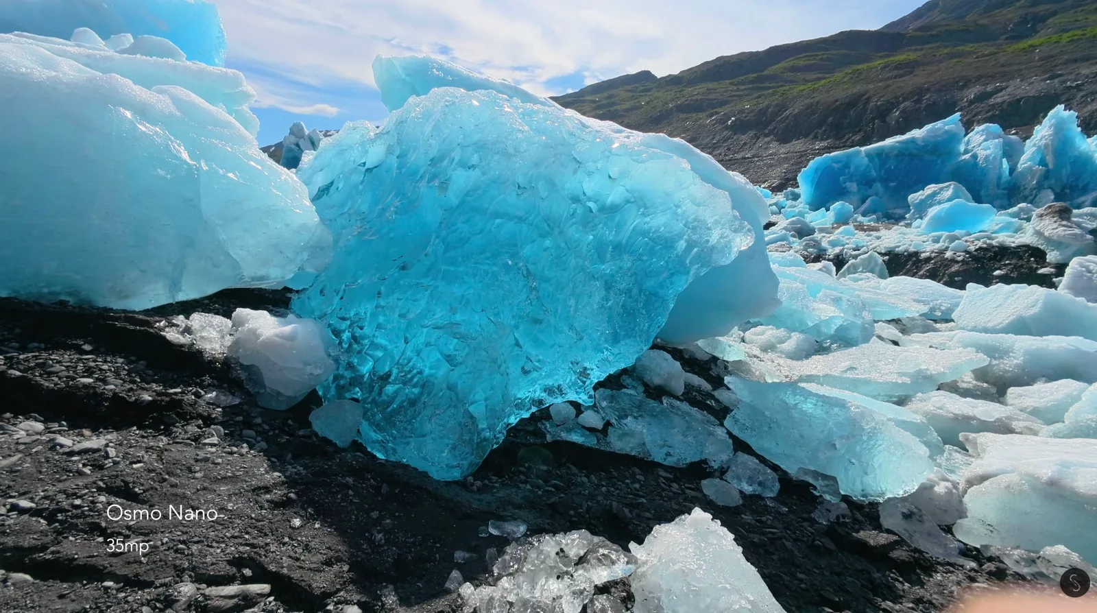 Translucent blue glacial ice chunks scattered across a dark gravel moraine under a partly cloudy sky, captured as a 35MP still with the DJI Osmo Nano