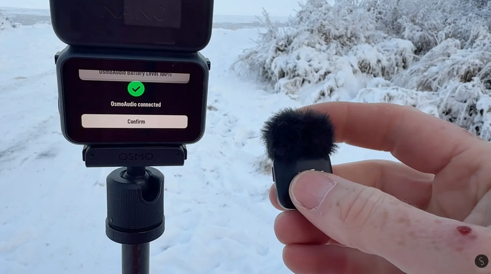 DJI Osmo Nano on a tripod in the snow showing an OsmoAudio connected confirmation screen, with a wireless microphone held in hand nearby