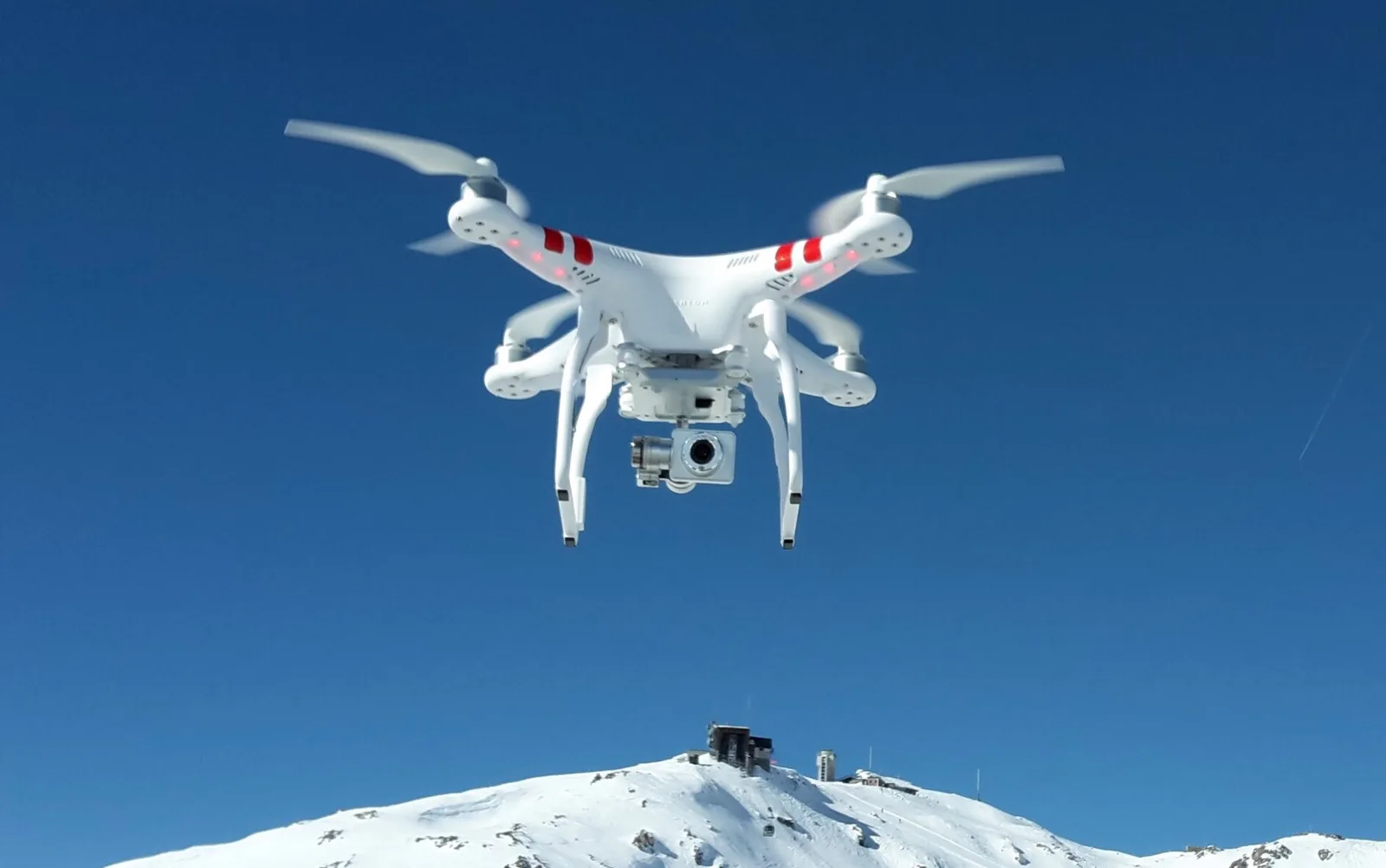 White DJI Phantom 2 Vision+ quadcopter hovering against a deep blue sky above a snow-covered mountain summit topped with a research station