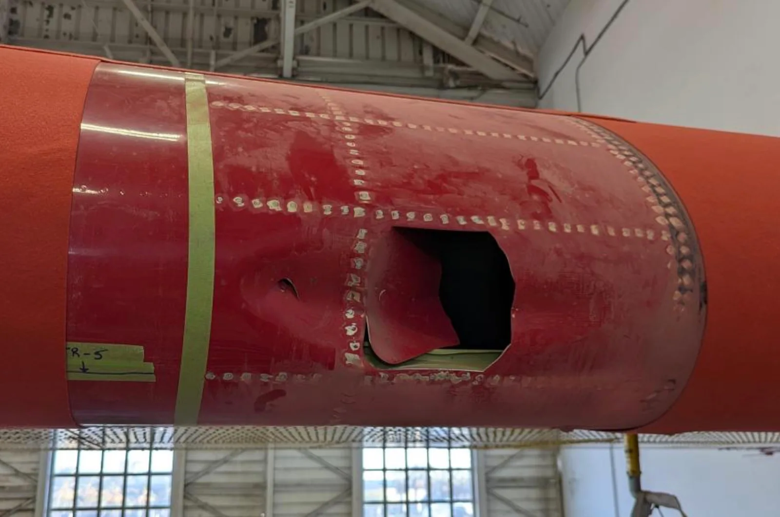 Jagged hole punched through the red leading edge of a Super Scooper firefighting aircraft wing, photographed in a hangar