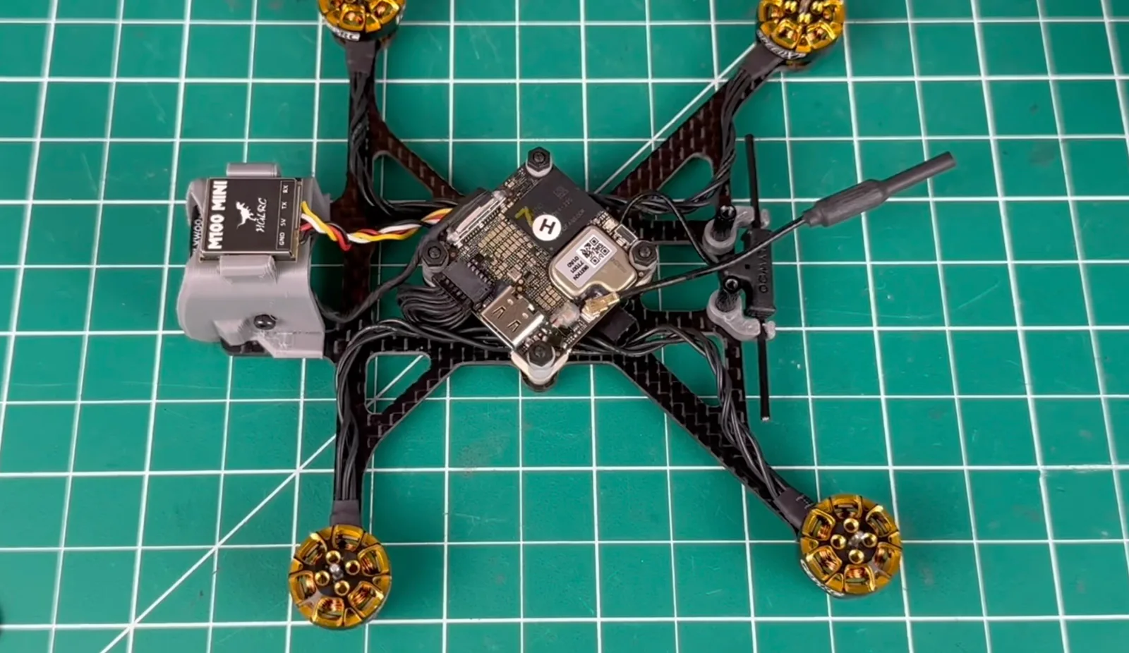Top-down view of an unfinished FPV freestyle quadcopter on a green cutting mat, showing its carbon fiber frame, gold-bell motors, exposed flight controller stack and a HGLRC M100 Mini GPS module.