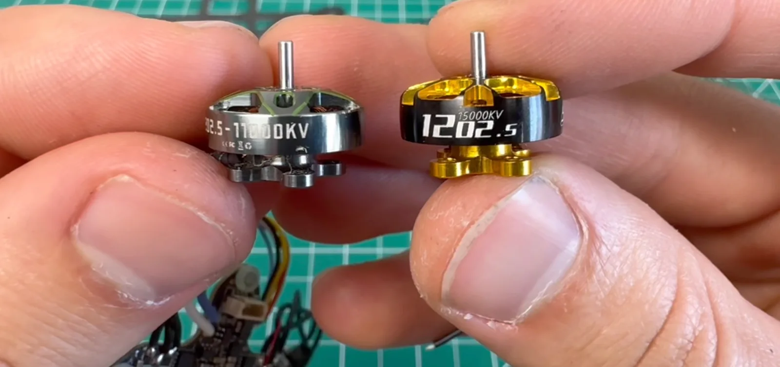 Two micro FPV drone brushless motors held side by side between fingers, a silver 1102.5 11000KV on the left and a gold-and-black 1202.5 15000KV on the right, above a green cutting mat.