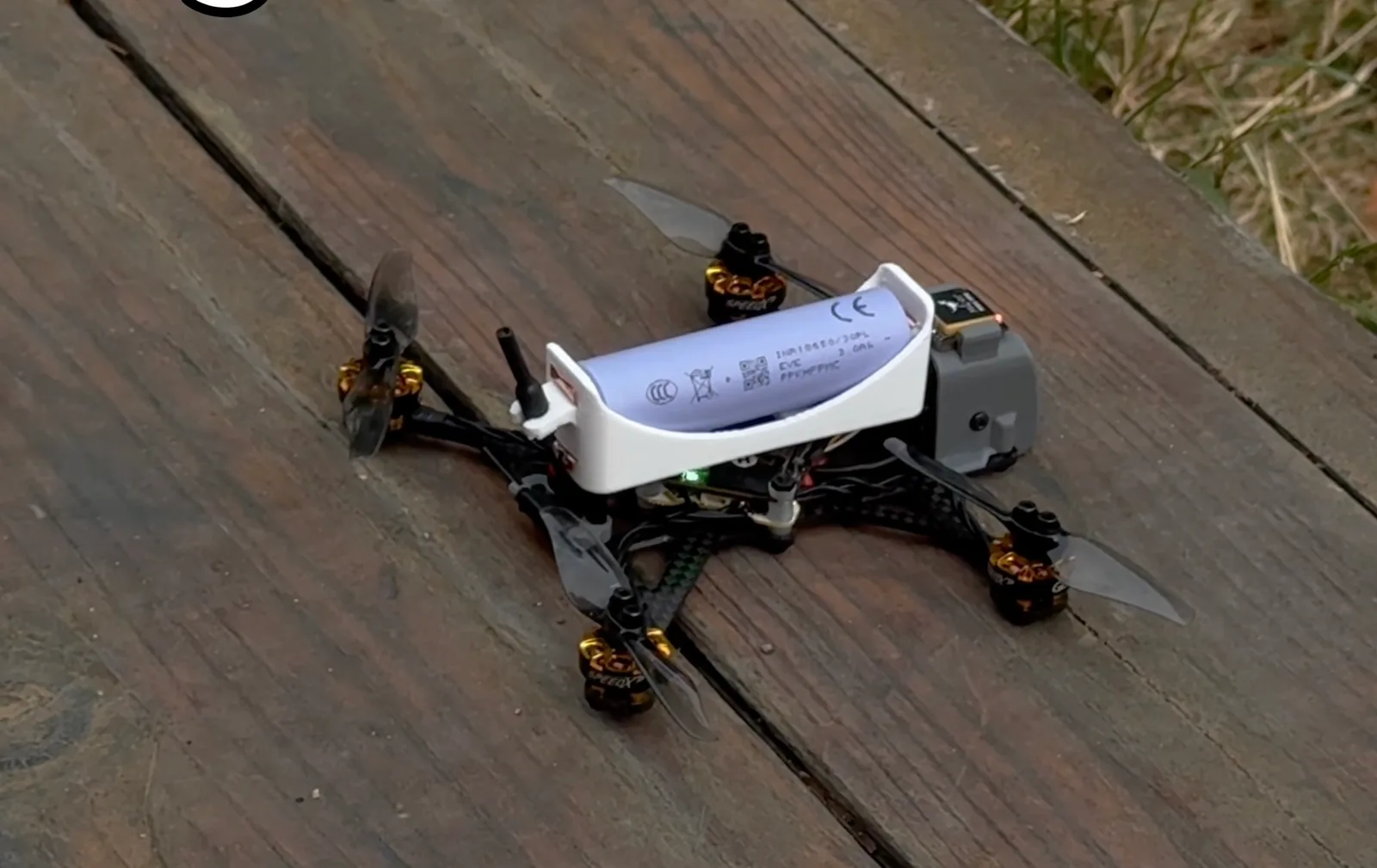Lightweight FPV racing quadcopter with carbon fiber arms and a single 18650 Li-ion cell resting on weathered wooden decking