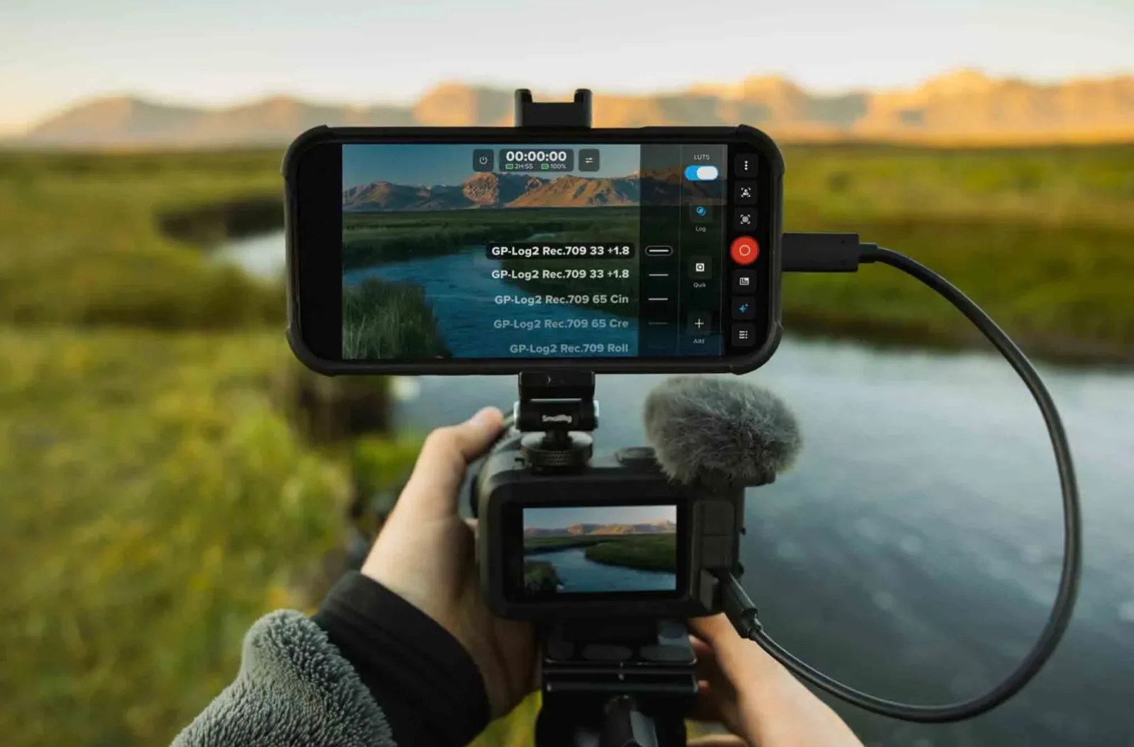 Hand-held GoPro MISSION 1 PRO ILS compact cinema camera with shotgun mic, tethered to a smartphone monitor showing GP-Log2 Rec.709 LUT options over a mountain river at sunrise