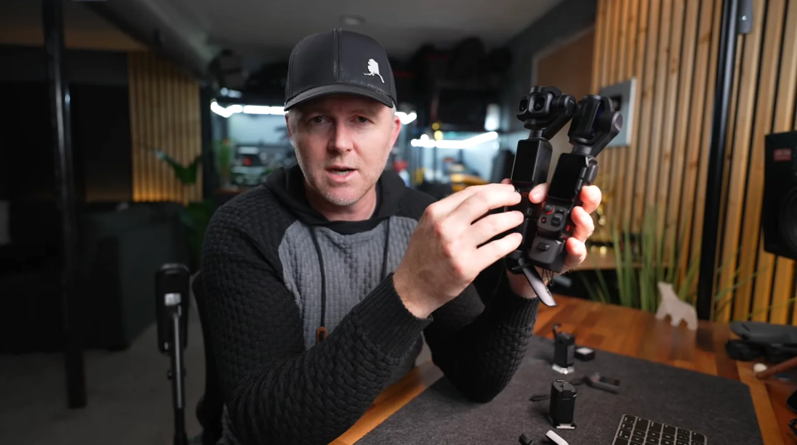 A reviewer holds up two pocket-sized handheld gimbal cameras side by side at a studio desk