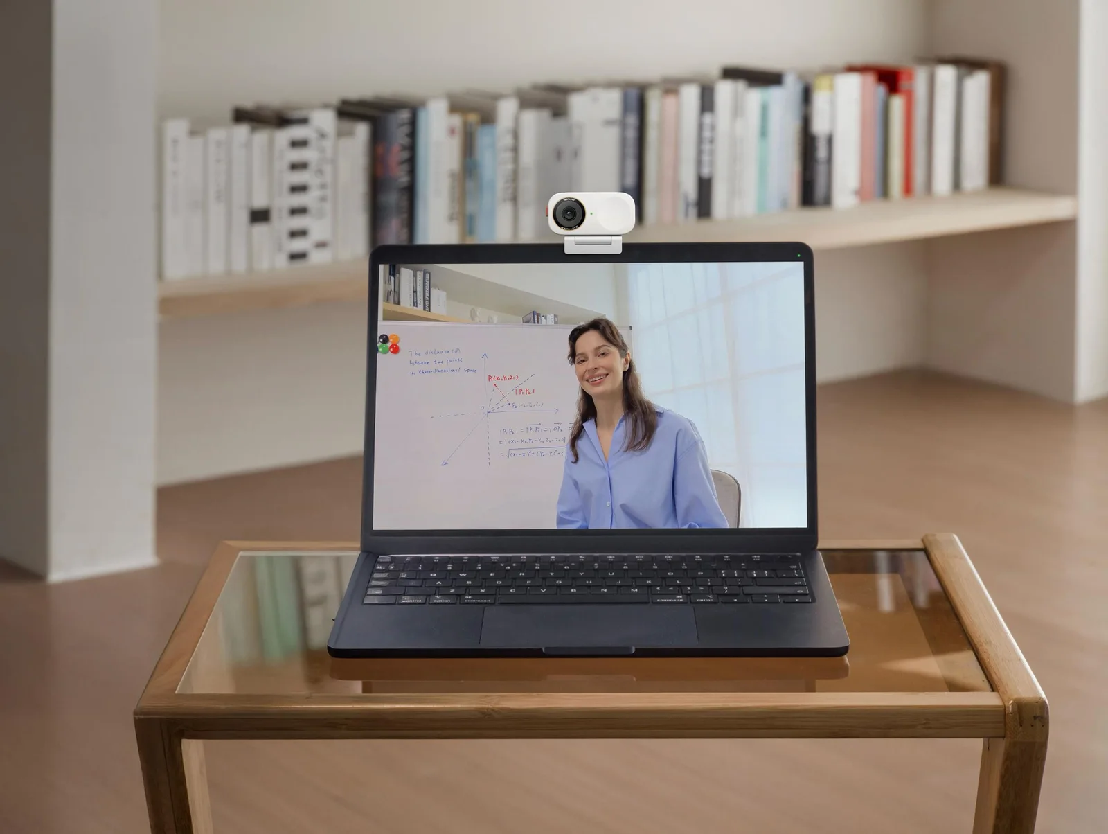 An Arctic White Insta360 webcam clipped to the top of a dark laptop on a wooden desk, with a video call on screen and a bookshelf behind.