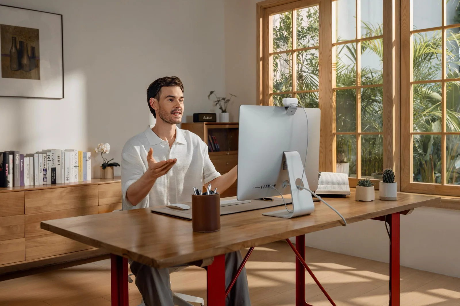 A white Insta360 webcam clipped to the top of an iMac display in a sunlit home office as a man gestures during a video call.