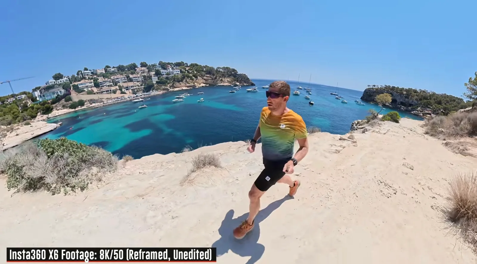 Trail runner in a gradient shirt and sunglasses running along a white limestone clifftop above a turquoise Mediterranean cove filled with anchored boats, captured in reframed Insta360 X6 8K footage