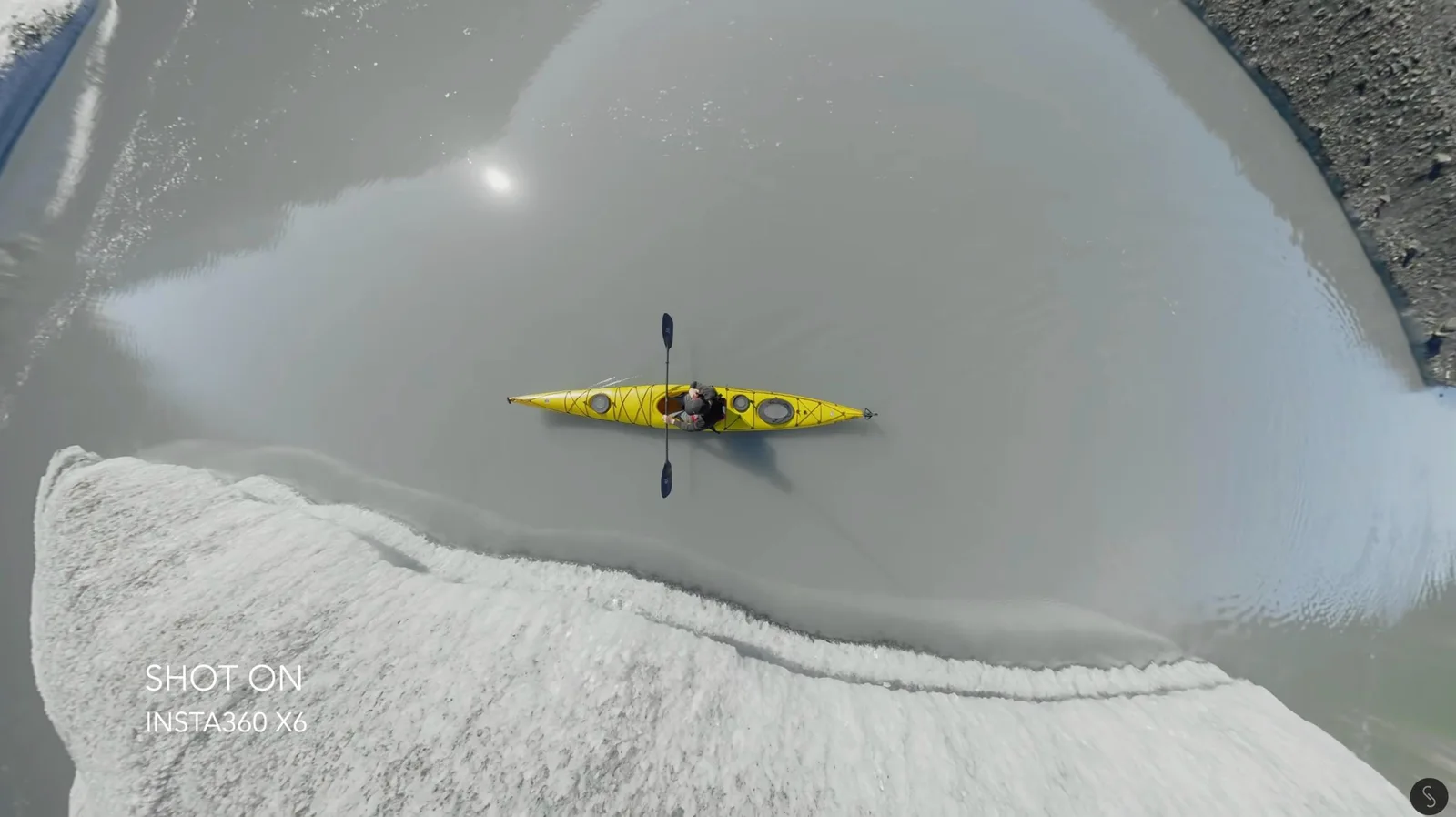 Overhead aerial view of a yellow sea kayak paddling across a still glacial lake beside a snow-covered ice shelf