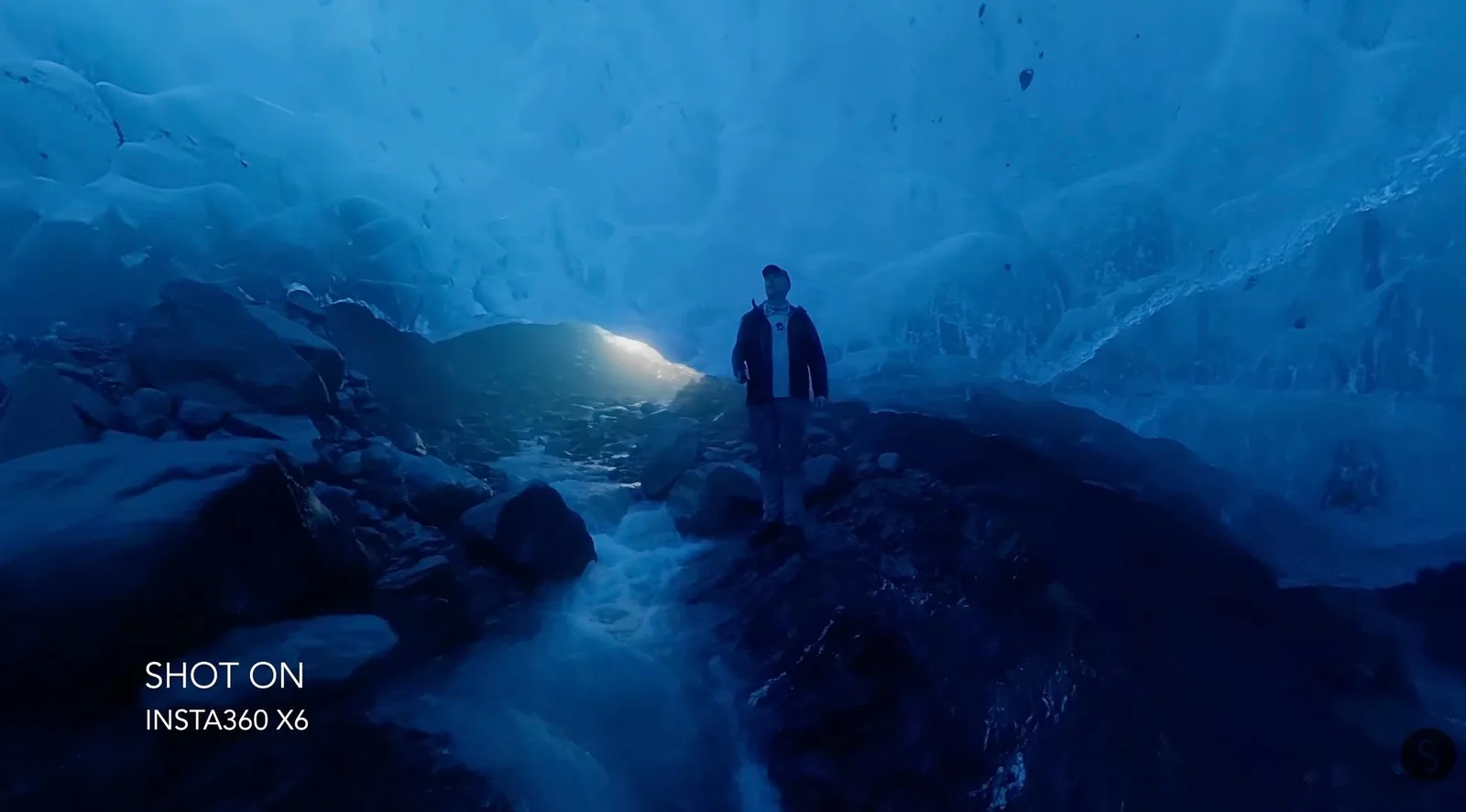 A person in a dark jacket and cap stands on rocks inside a deep blue glacier ice cave, with a meltwater stream running past and daylight glowing through the cave mouth behind them.
