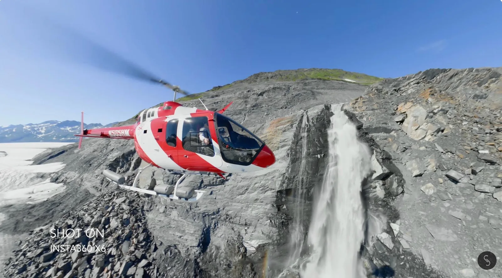 Red and white Bell helicopter hovering beside a waterfall on a rocky Alaskan mountainside, captured with a 360 camera