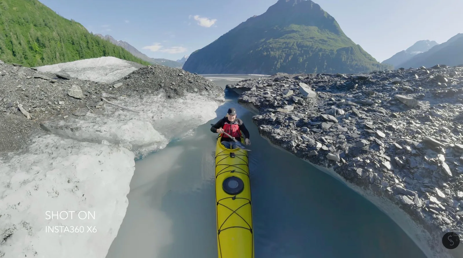 Kayaker paddling a yellow sea kayak through a narrow glacial meltwater channel between ice and rock moraine, with green mountains rising ahead in Alaska
