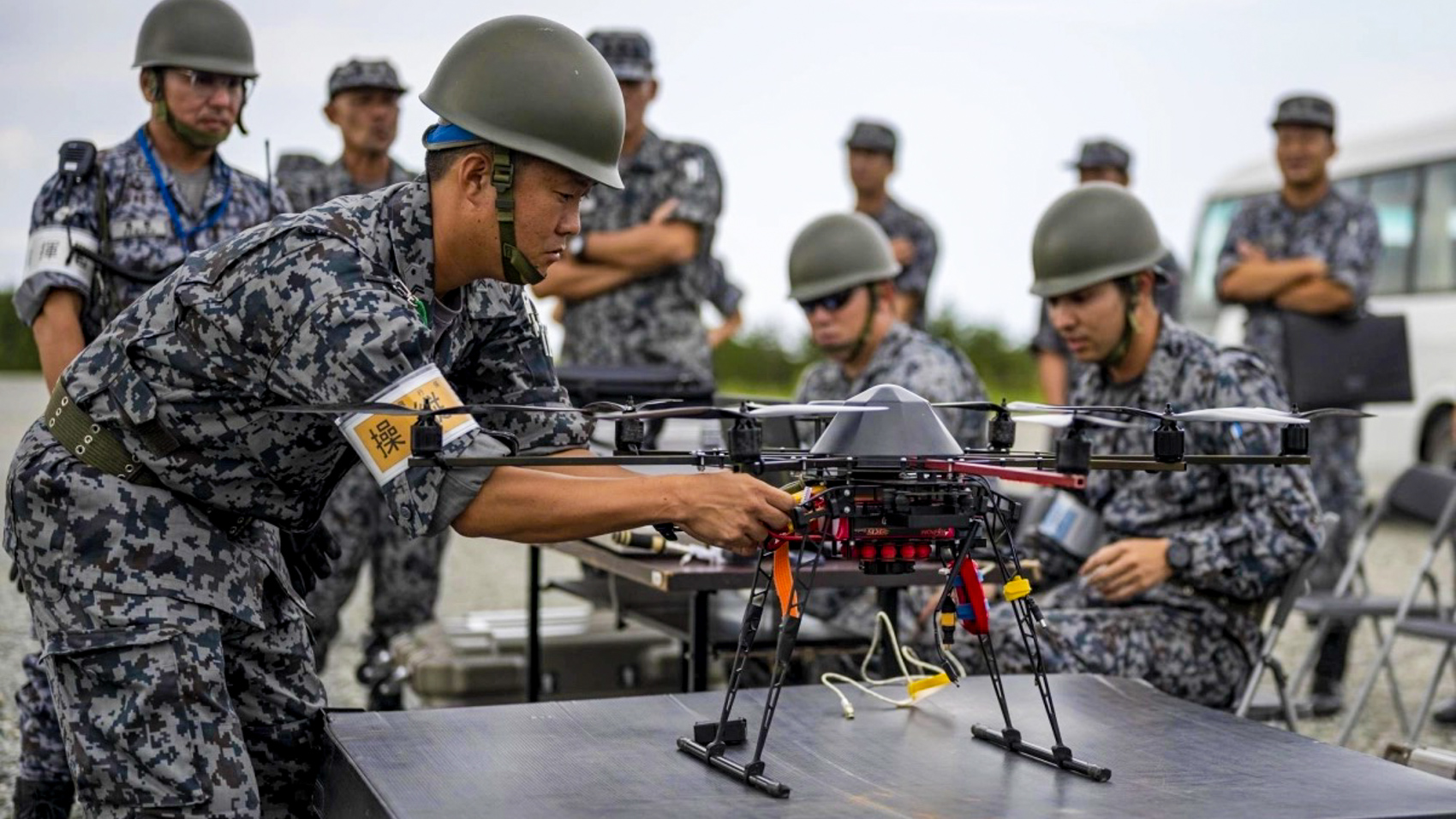 A Japanese airman in blue digital camouflage and a helmet prepares a black multirotor drone on a table while other service members watch.