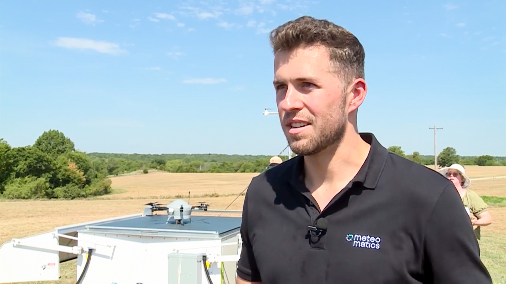 A man in a black Meteomatics polo shirt speaks to an interviewer in an Oklahoma field, with a drone parked on a white mobile ground station behind him.