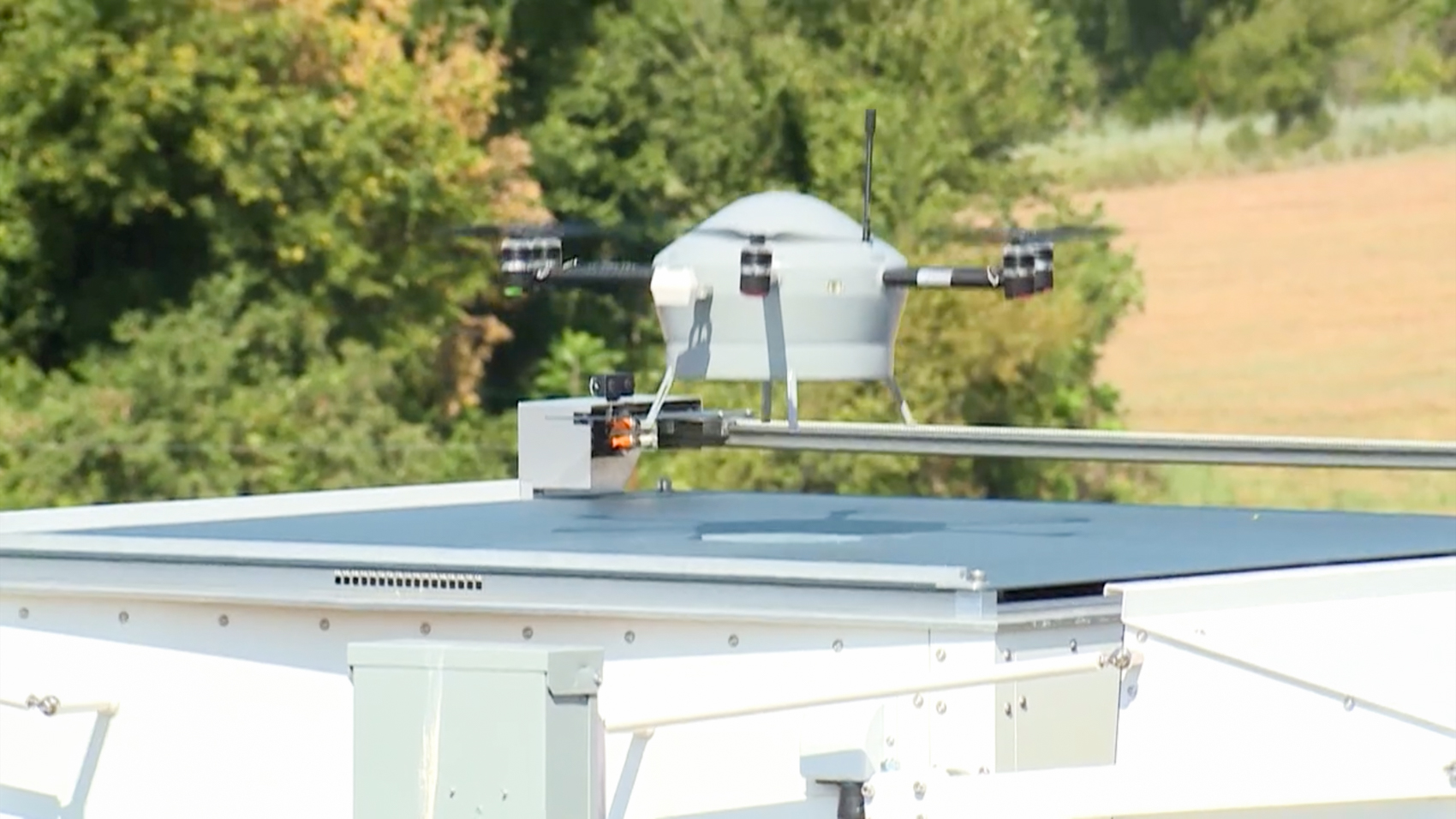 A white Meteodrone quadcopter lifts off from the flat roof of an automated white ground station, with green trees and a farm field behind it.