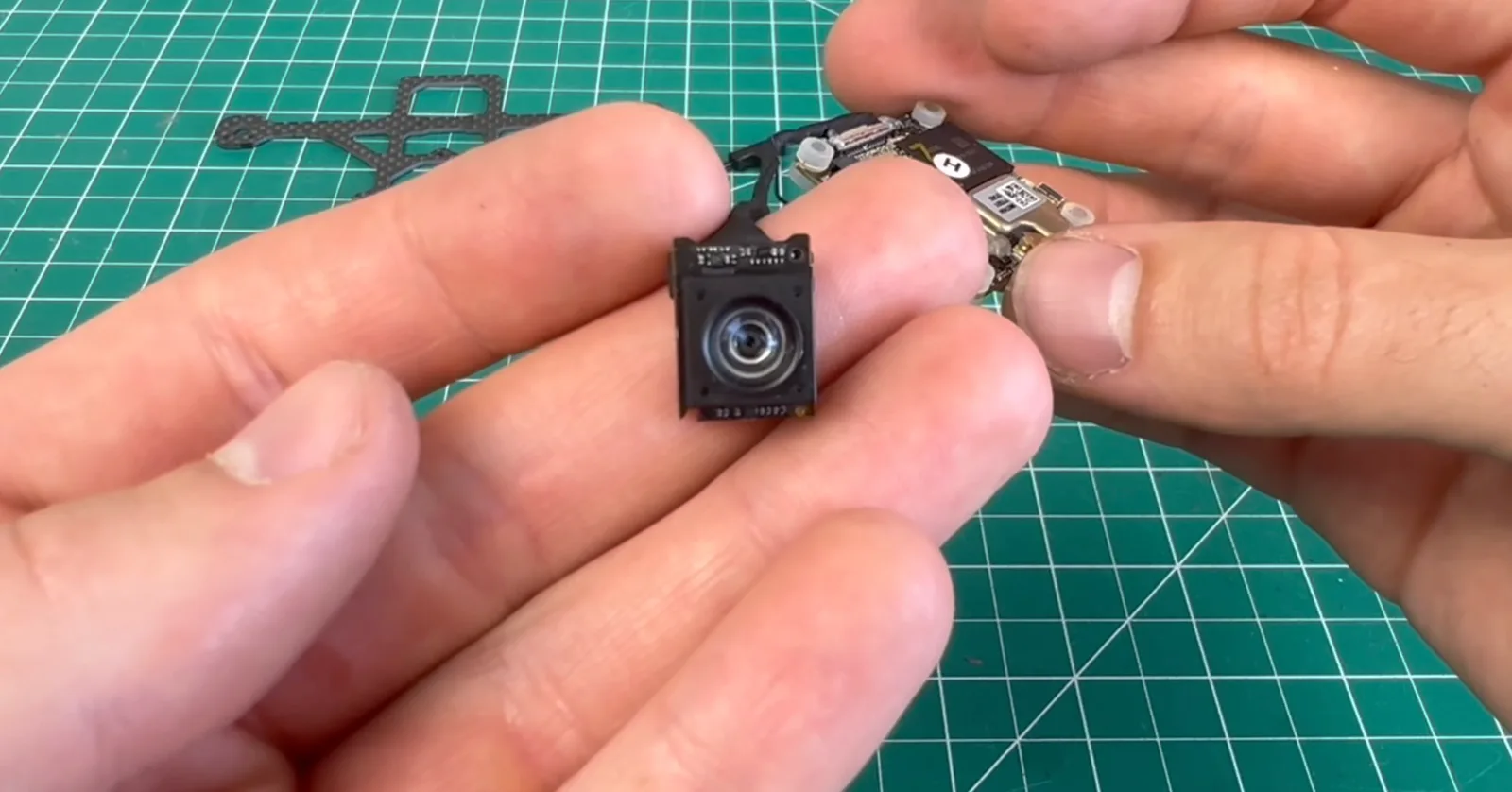 Hands holding a tiny FPV drone camera module and its flight controller board over a green cutting mat