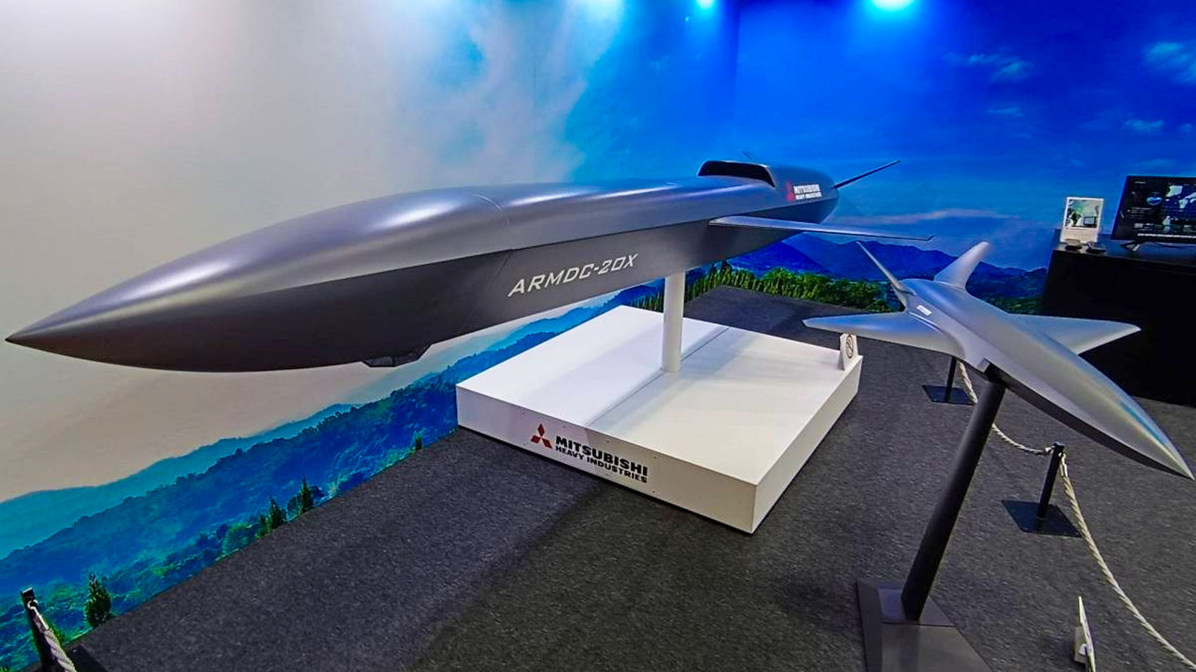 A large dark grey uncrewed aircraft model labeled ARMDC-20X sits on a Mitsubishi Heavy Industries display plinth beside a smaller silver airframe model.