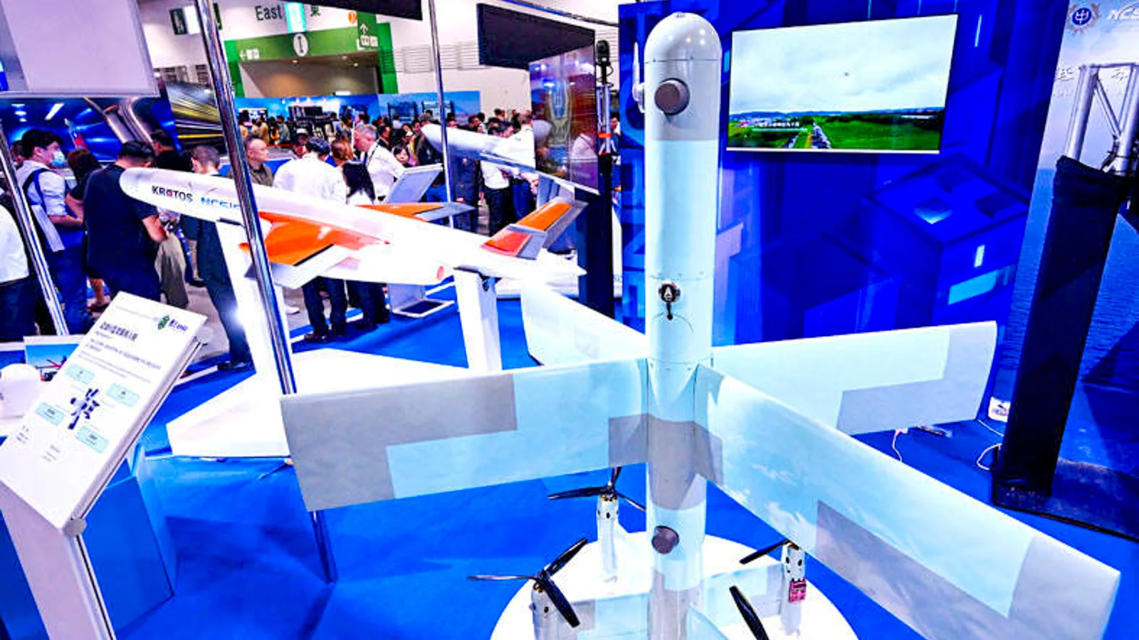 A Mighty Hornet III X-wing drone displayed on a rotating stand on a crowded Taiwanese defence exhibition floor, with other NCSIST aircraft and visitors behind it
