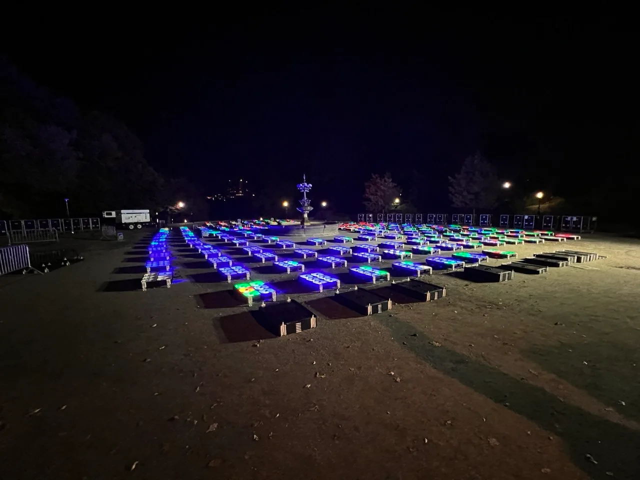 Rows of illuminated drone launch pads glowing blue and green on a dirt field in Central Park at night, staged before a Nova Sky Stories drone light show