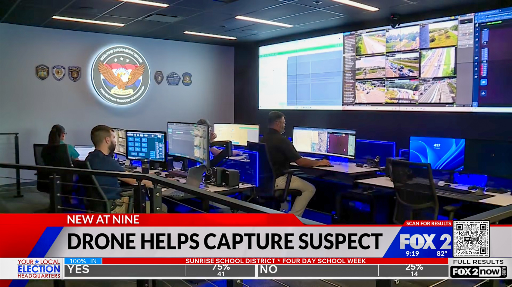 Operators at rows of monitors in a darkened Real Time Information Center, with a wall of live camera and drone feeds and the O'Fallon police crest.
