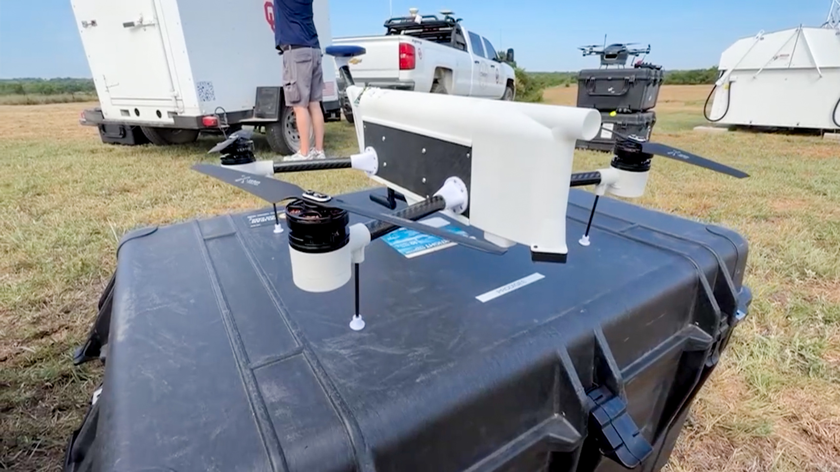 A white research drone with a tubular sensor housing rests on a hard case in a grass field, with a university trailer, pickup truck and a second drone staged behind it.