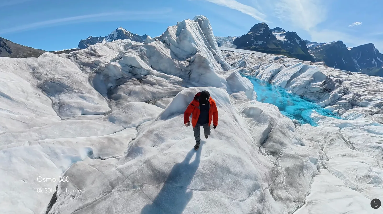 Hiker in an orange jacket walking along a glacier ridge with turquoise meltwater and snow-capped peaks behind, captured on a DJI Osmo 360