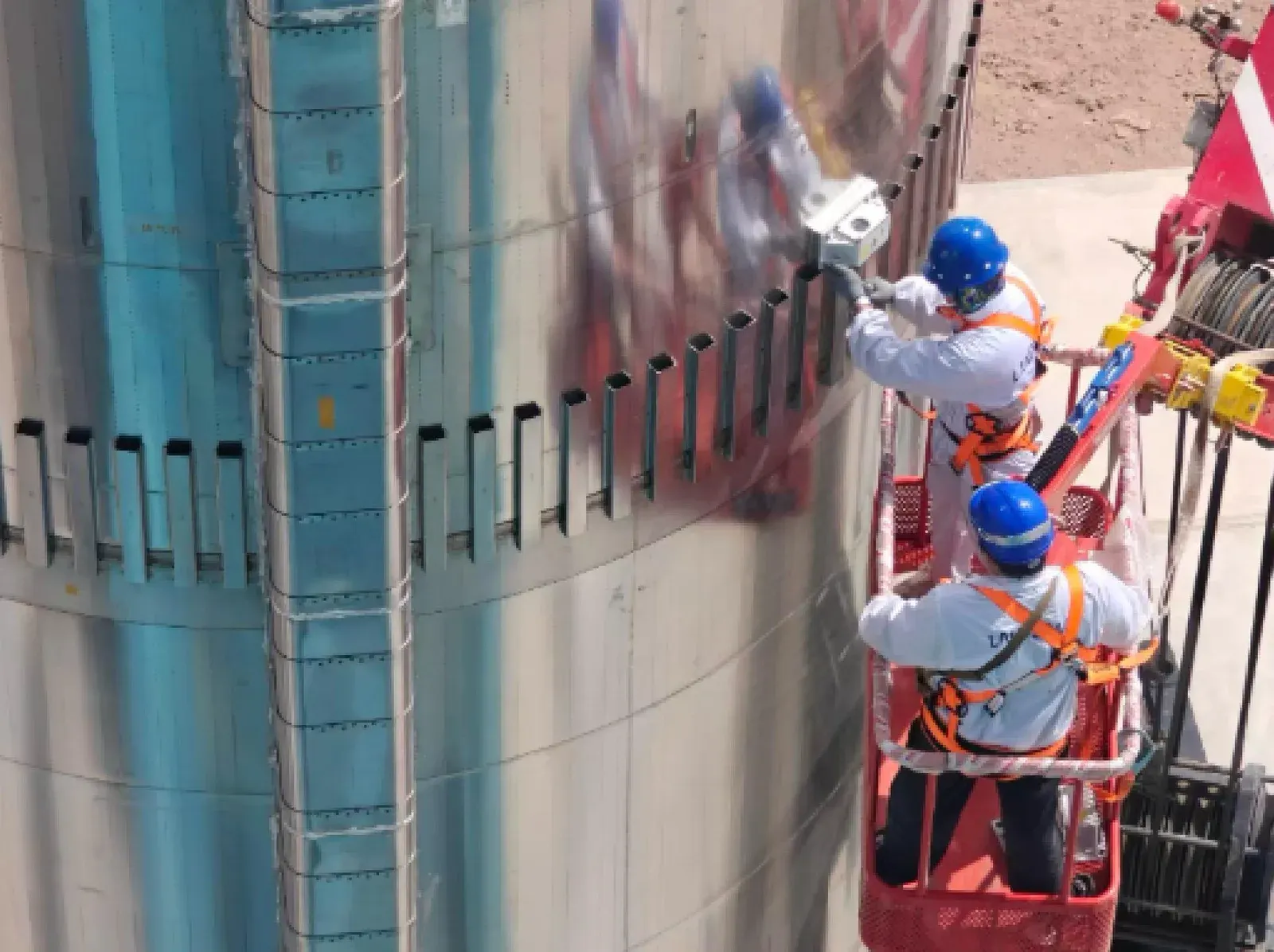 Two technicians in white coveralls and blue hard hats stand in a red cherry-picker basket, bolting a white action camera to the side of a rocket booster stage.