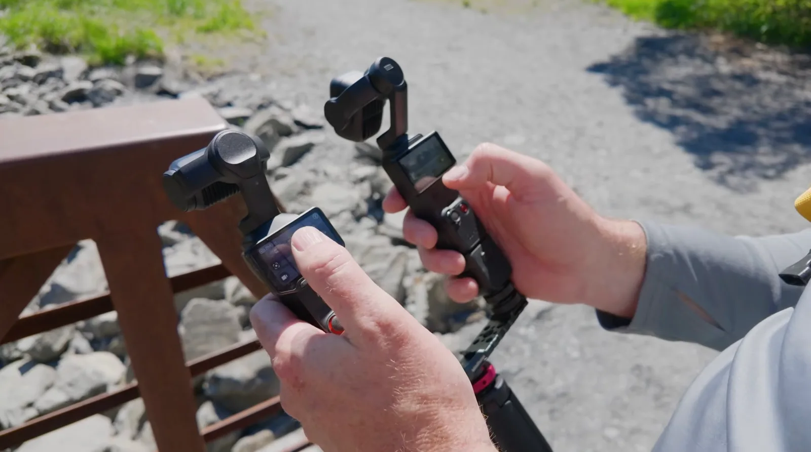 Two handheld pocket gimbal cameras held side by side outdoors, one with its flip-out touchscreen open showing the camera interface