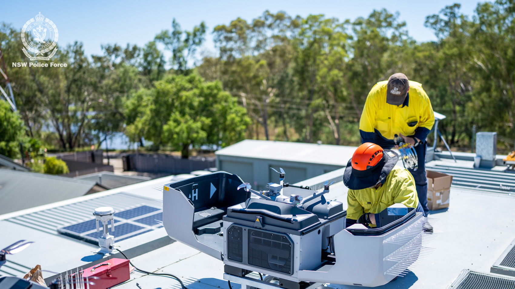 Two technicians in high-visibility clothing install a white drone docking station on a metal police station roof.