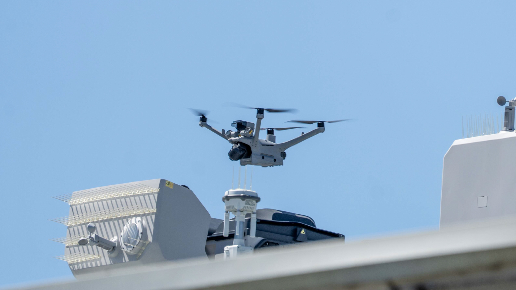 A police drone flies above rooftop equipment including its docking station and antennas.