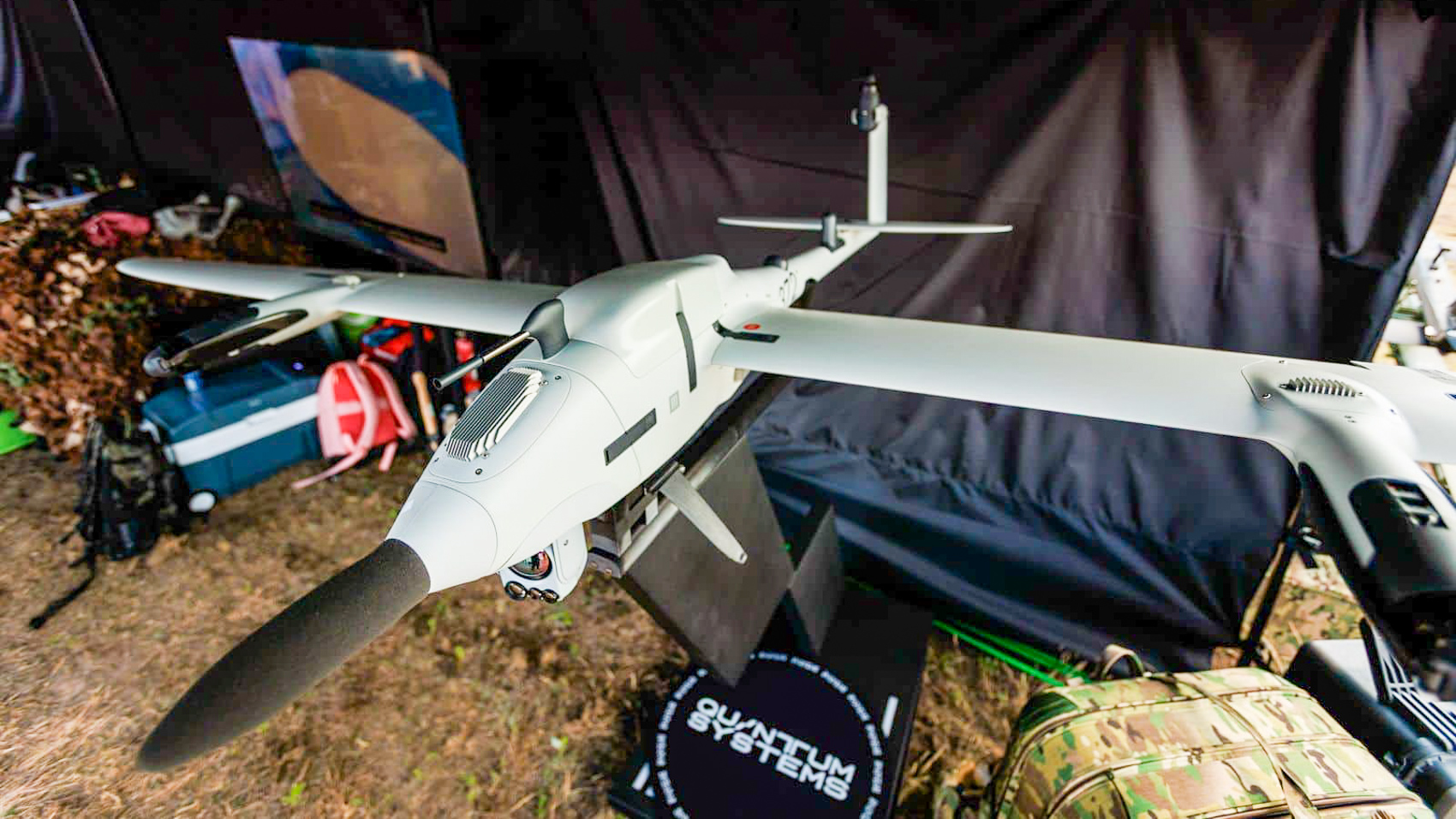 A Vector drone sits under a camouflage net at a field position, its tail boom and sensor mast visible beside stacked equipment cases.