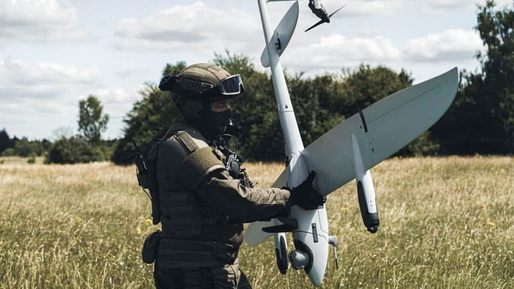 A soldier in full tactical gear holds a grey Vector fixed-wing drone upright by its fuselage in a dry summer field.
