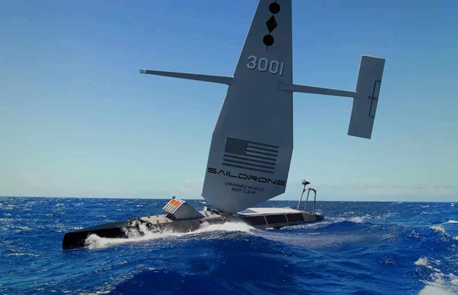 Saildrone uncrewed surface vessel hull number 3001 sailing through deep blue open ocean under a clear sky, its tall gray wing sail marked with a US flag and the words UNMANNED VEHICLE KEEP CLEAR