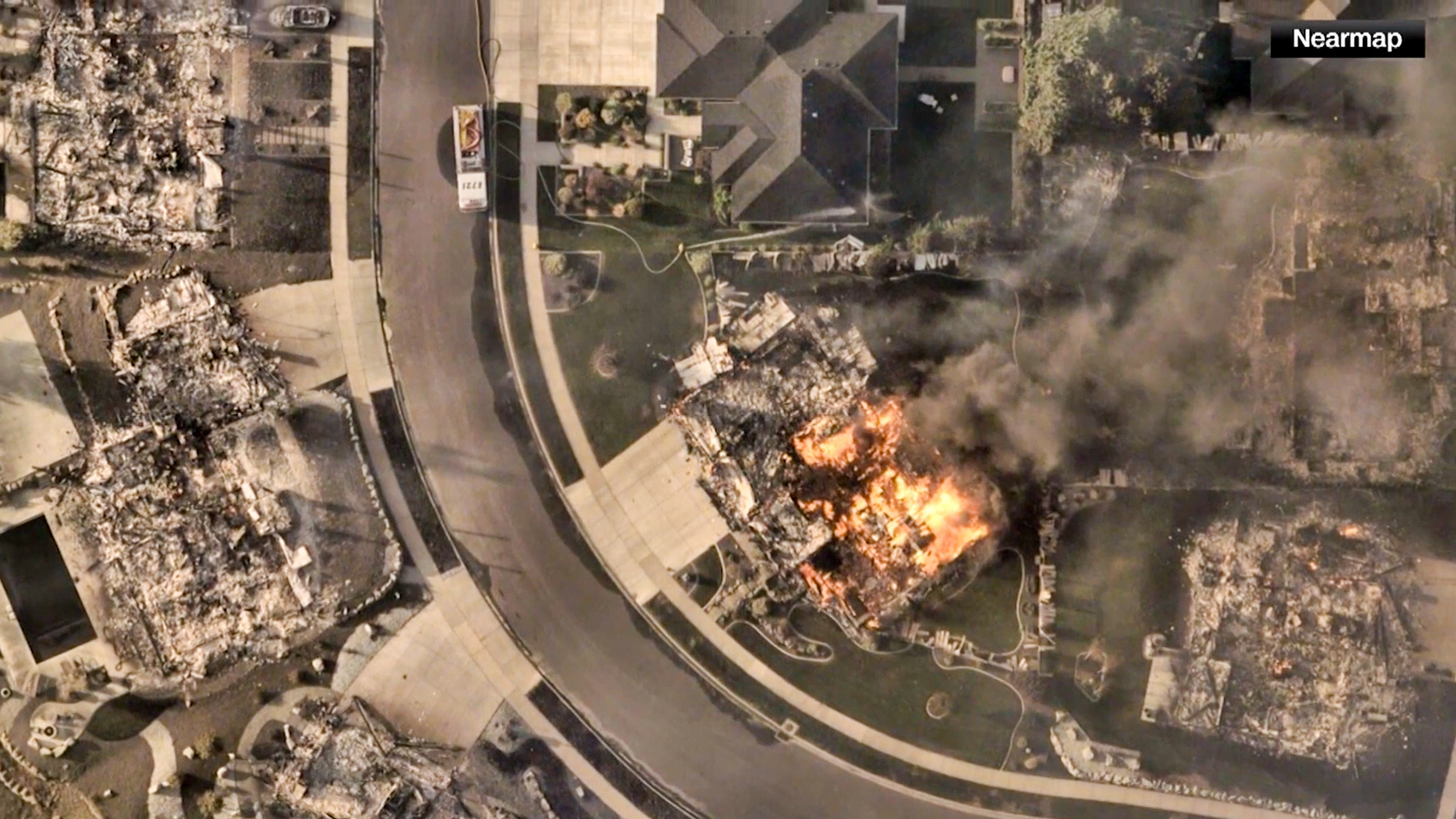 Overhead aerial image of a cul-de-sac where several homes are burned to their foundations and one house is engulfed in open flames and smoke.
