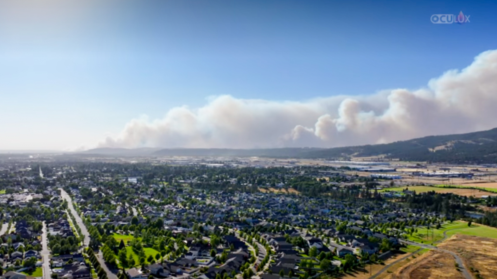 Aerial view of massive smoke plumes rising from wildfires on forested ridges beyond a suburban neighborhood in the Spokane Valley.