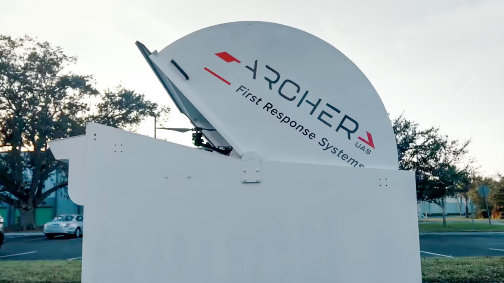 A white Archer First Response Systems drone dock with its domed lid opening in a parking lot lined with trees