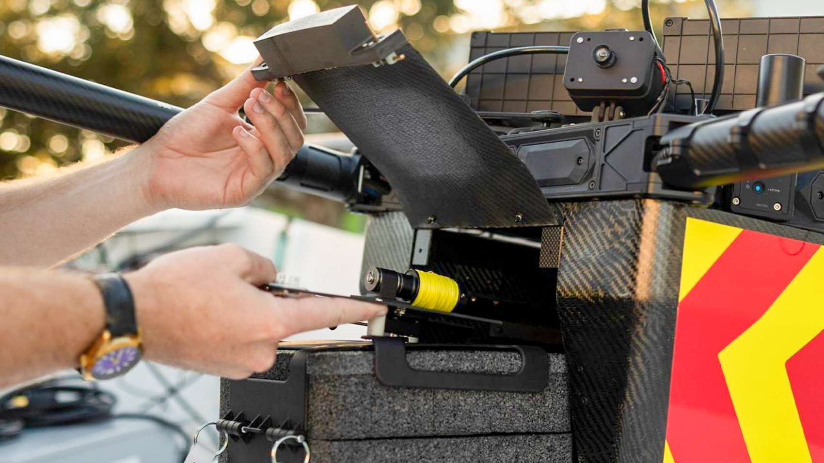 Hands loading a tethered payload spool and insulated supply box into a delivery drone marked with red and yellow chevrons