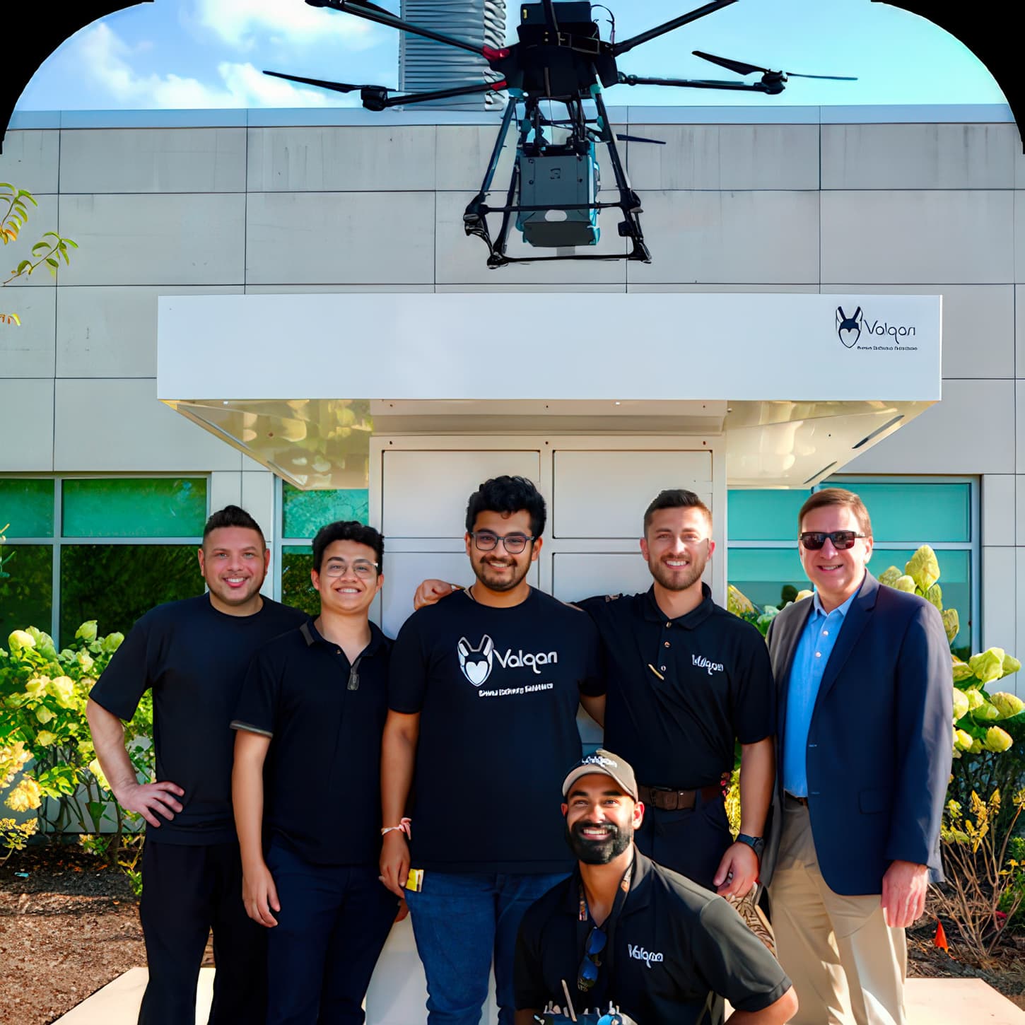 The Valqari team poses in front of a smart delivery station as a delivery drone hovers overhead