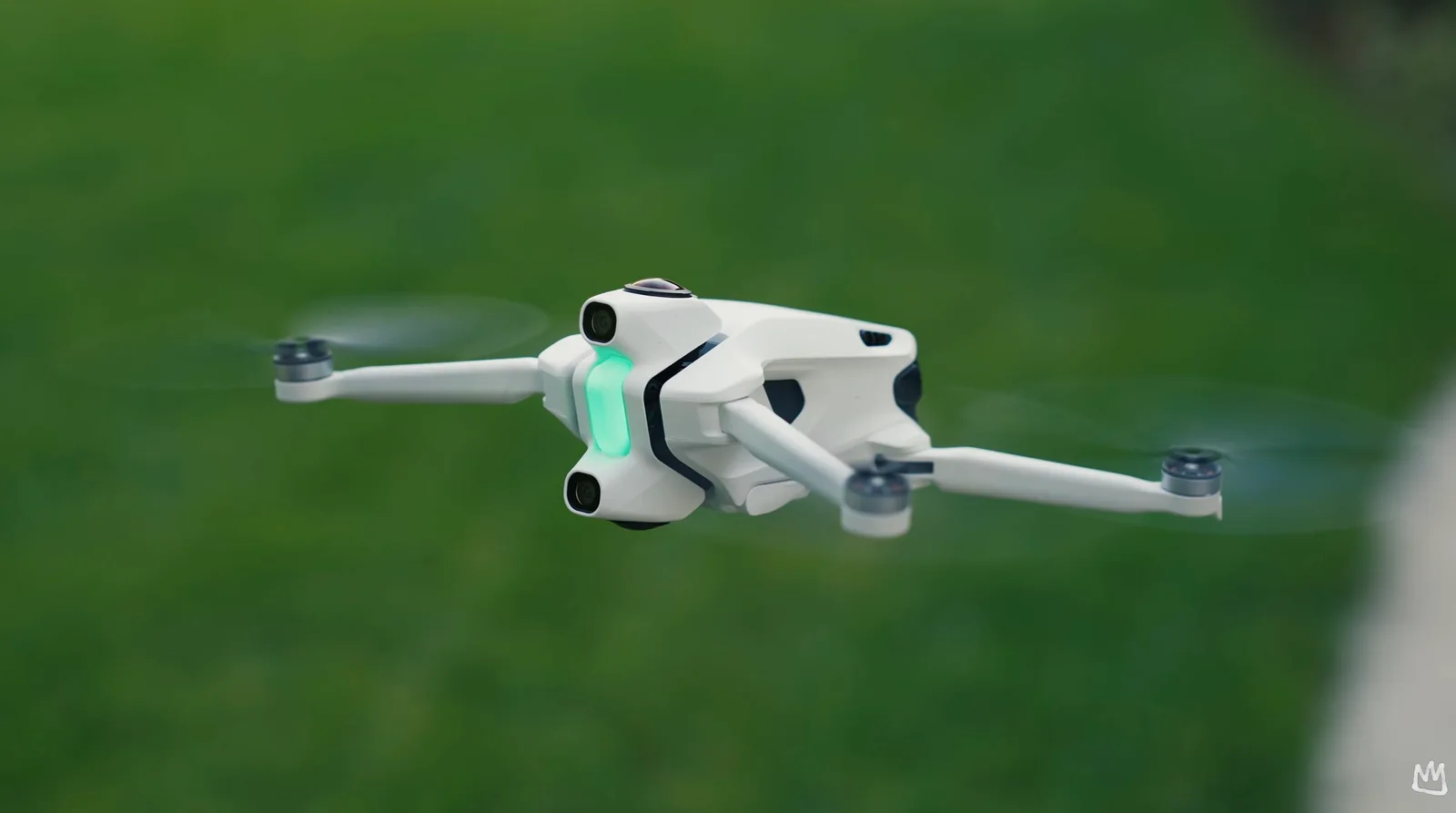 White compact folding drone hovering above a grass field with its propellers spinning and green status light glowing