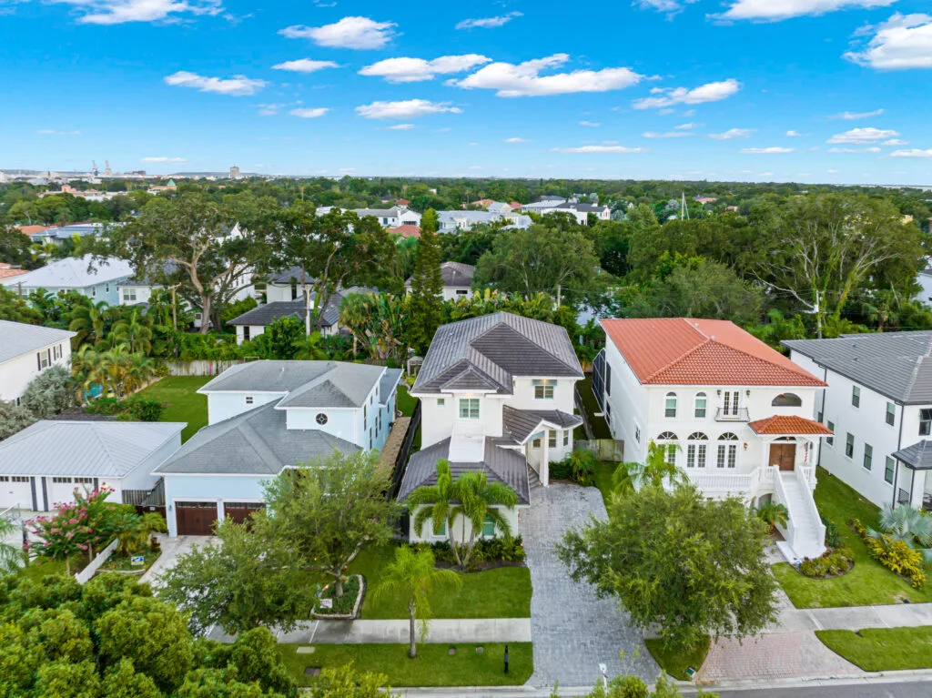 Aerial drone photo of a two-story white home with a gray roof and paver driveway between neighboring houses in a tree-lined Tampa neighborhood