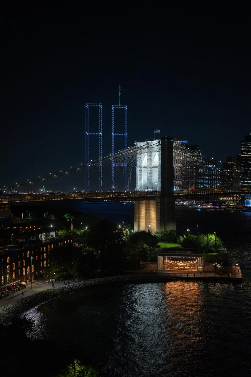 Drone light show recreating the Twin Towers in light beams behind the Brooklyn Bridge over New York Harbor at night