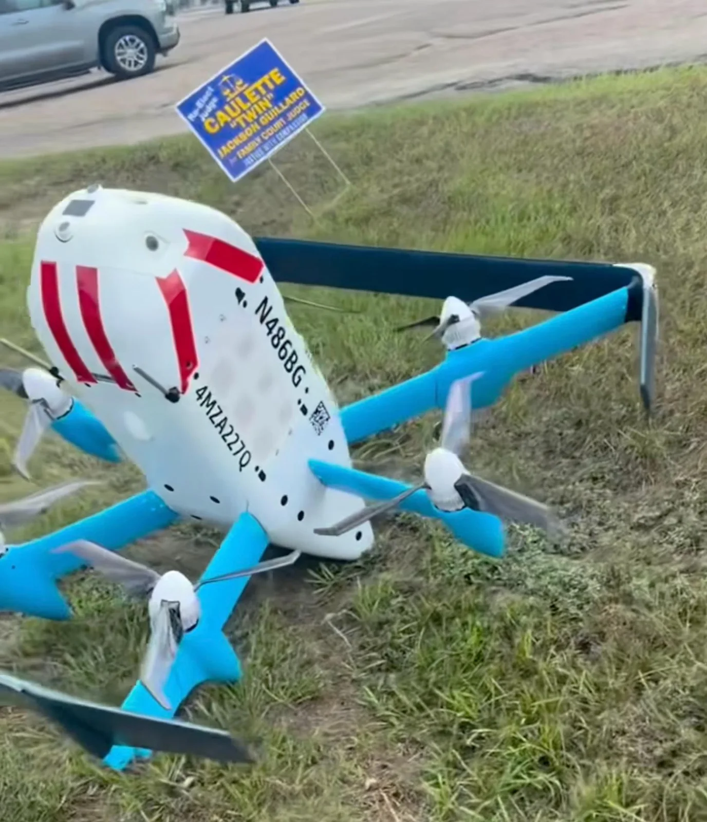 Amazon MK30 delivery drone on the grass shoulder of Florida Boulevard in Baton Rouge with a campaign yard sign and passing traffic behind it